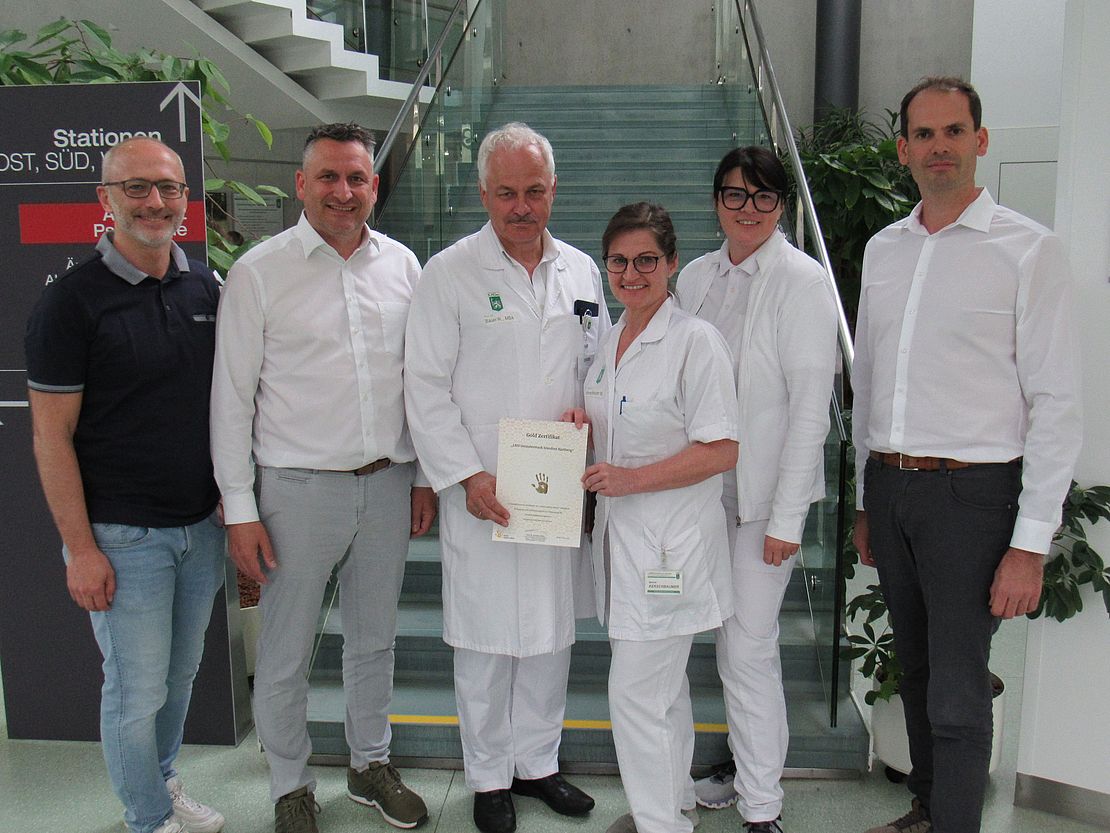 Gruppenfoto vor einer Stiege im Krankenhaus, eine Person hÀlt Zertifikat